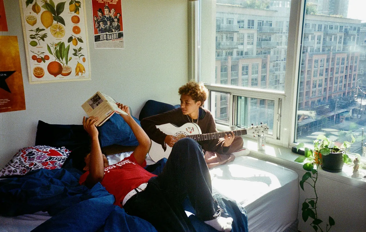 Dos estudiantes compartiendo habitación en una residencia: uno leyendo tumbado en la cama y otro tocando la guitarra junto a la ventana.